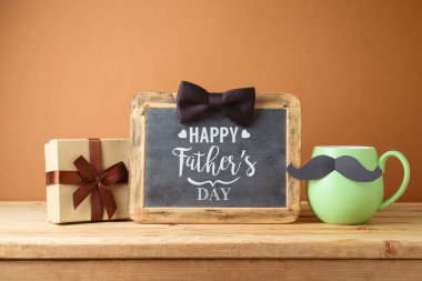 Happy Fathers day greeting card with green coffee cup, mustache, gift box and chalkboard on wooden table