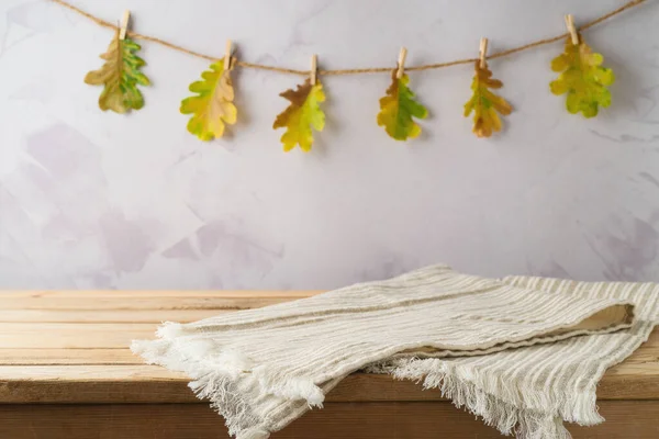 Empty wooden table with modern tablecloth over autumn leaves garland background. Autumn kitchen mock up for design and product display