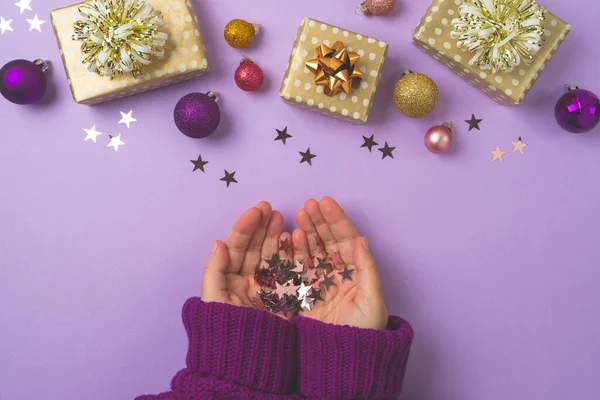 Christmas concept with gift box, woman hands and decorations on purple background. First person view. Top view, flat lay