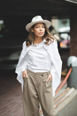 Beyaz pantolonlu, beyaz pamuklu bluzlu şık bir kız duvara poz veriyor. Şapkalı bir kadının fotoğrafı..