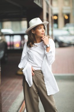 Beyaz pantolonlu, beyaz pamuklu bluzlu şık bir kız duvara poz veriyor. Şapkalı bir kadının fotoğrafı..