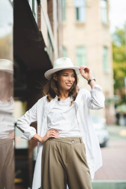 Beyaz pantolonlu, beyaz pamuklu bluzlu şık bir kız duvara poz veriyor. Şapkalı bir kadının fotoğrafı..
