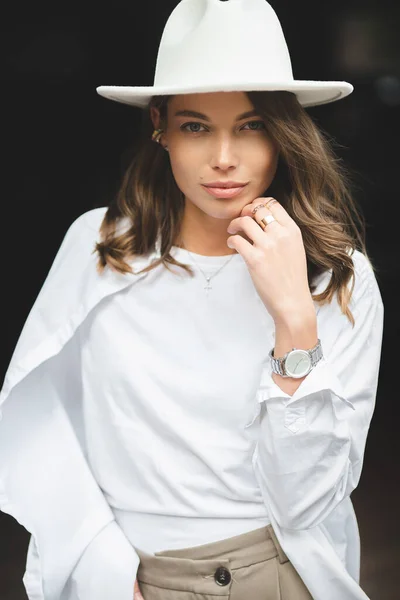 穿着白色长裤和白色棉衣的时髦女孩靠墙摆姿势 一个戴帽子的女人的照片 — 图库照片