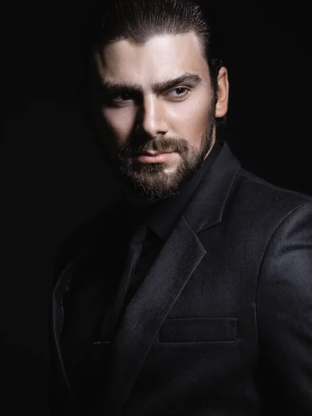 Young white Caucasian man on a black background in a black jacket, shirt and black tie looks out from under his forehead. Strict male portrait. The guy in the classic black suit.