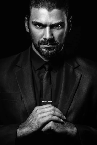 Black and white male portrait. A man in a black classic suit in a black shirt and tie over a black background. Dark-haired young man.