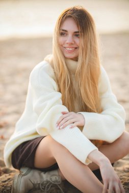 Çekici bir sarışının harika bir gülümsemesi olan portresi. Beyaz kazaklı bir kız nehir kıyısında dinleniyor.