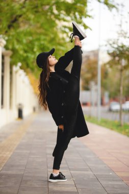 Girl gymnast with a raised leg above her head on a city street. An attractive girl in a coat and a baseball cap is engaged in gymnastics on the street