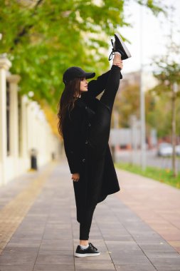 Girl gymnast with a raised leg above her head on a city street. An attractive girl in a coat and a baseball cap is engaged in gymnastics on the street