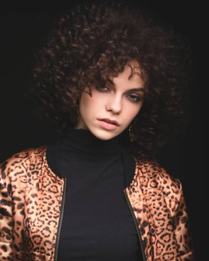 Afrikalı kıvırcık saçlı beyaz bir kızın modaya uygun portresi. Siyah arka planda leopar desenli ceketli bir kız.