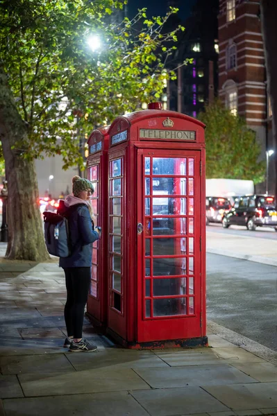 2021年11月2日 一位年轻女子 夜晚背着背包走出传统的英国红色电话亭 — 图库照片