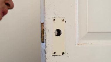 Bir adam kapı kilidi namlusundaki eski beyaz bir iç kapının döküntülerini temizliyor.