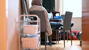 Düşük açılı kayma görüntüsü, masadaki dizüstü bilgisayarda çalışan kapüşonlu sabahlıklı bir kadını gösteriyor..