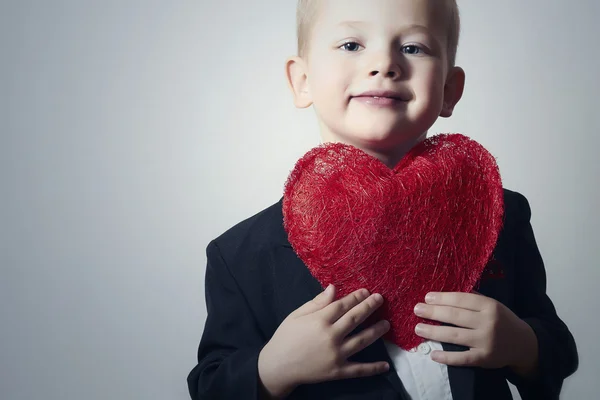 Niño sonriente con corazón rojo. Niño de cuatro años con símbolo del corazón. Niño encantador en traje negro Día de San Valentín —  Fotos de Stock