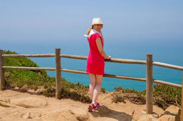 Jeune voyageuse avec chapeau blanc et robe rouge restant près de la clôture en bois à la falaise de Cabo da Roca Cape Roca — Photo