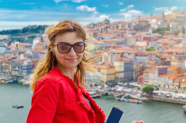 Mujer joven viajera con gafas de sol y chaqueta roja mirando a la cámara, sonriendo y posando frente a la ciudad de Porto Oporto — Foto de Stock