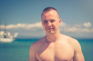 Yakından çekilmiş genç kaslı üstsüz erkek vücut geliştiren atletik vücutlu adam. Kameraya bakıyor ve gülümsüyor. Güneşli yaz gününde Halkidiki 'de Toroneos Kolpos Körfezi' nin turkuaz suyu.