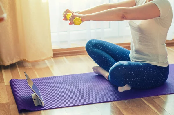 Woman doing workout sports at home, raising dumbbells with straight arms, sitting on floor mat crossing legs and looking at tablet screen in living room, close-up view, sport and recreation concept
