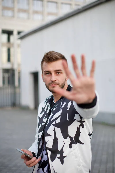 A handsome man in a white and black suit hides from the paparazzi and sticks out his hand. Guy with stubble and a model hairstyle in a fashionable suit sticks out his hand so as not to be filmed.