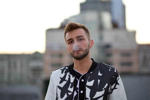 A handsome man in a white and black suit exhales the smoke of a cigarette. A guy with stubble and a model hairstyle in a fashionable suit and smoke from a cigarette.