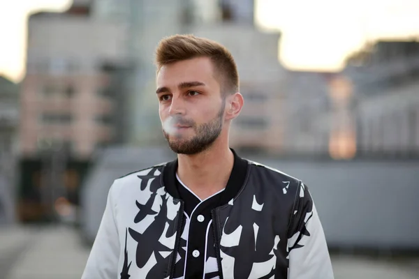 A handsome man in a white and black suit exhales the smoke of a cigarette. A guy with stubble and a model hairstyle in a fashionable suit and smoke from a cigarette.