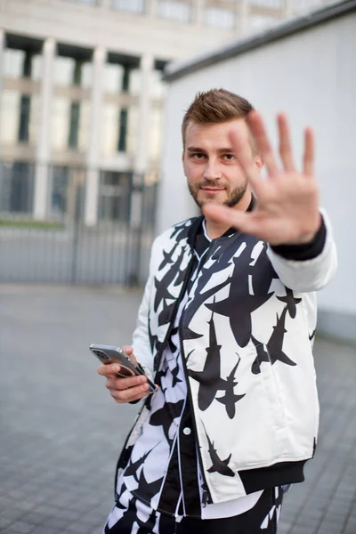 Handsome man in a white and black suit hides from the paparazzi and sticks out his hand. Guy with stubble and a model hairstyle in a fashionable suit sticks out his hand so as not to be filmed