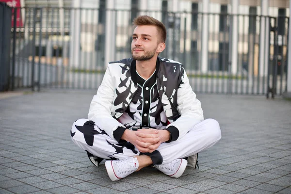 Handsome man in a white and black suit with a playful look. A guy with stubble and a model hairstyle in a trendy suit sits on the pavement