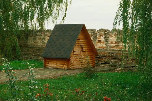 Wooden ancient well with a tiled roof. An old well in the park and a path to it. A brick wall can be seen in the background.