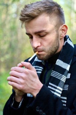 A handsome guy in a black coat and black and white scarf smokes a cigarette. Smoking - a guy of model appearance in a coat and a scarf with a cigarette.