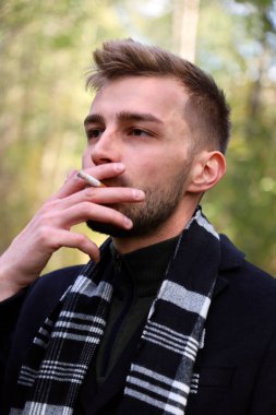 A handsome guy in a black coat and black and white scarf smokes a cigarette. Smoking - a guy of model appearance in a coat and a scarf with a cigarette.