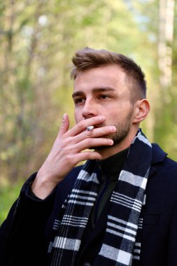 A handsome guy in a black coat and black and white scarf smokes a cigarette. Smoking - a guy of model appearance in a coat and a scarf with a cigarette.