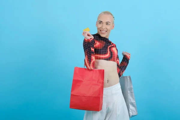 Junge nicht binäre Person, die eine Kreditkarte und Einkaufstüten in der Hand hält, während sie auf einem isolierten Hintergrund posiert. — Stockfoto