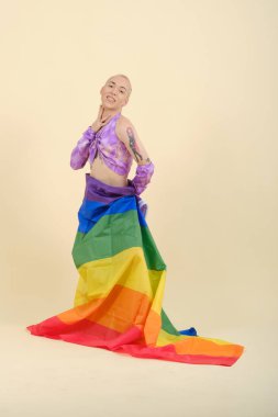 İkili olmayan kişi kameraya bakıyor ve beline bağlı bir gökkuşağı bayrağıyla poz verirken gülümsüyor..