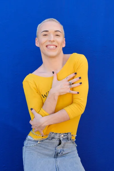 Young non-binary person looking at the camera and smile while posing outdoors over a blue background. — Stock Photo, Image