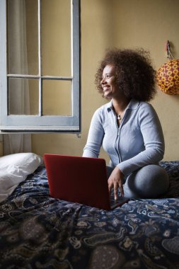 Evdeki bir yatakta rahatça otururken bilgisayarını kullanan gülümseyen kadın..