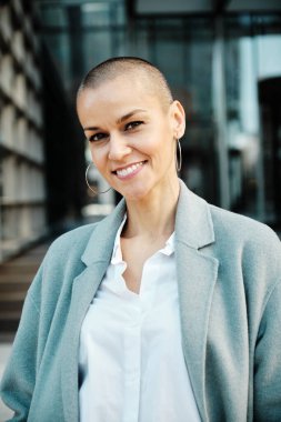 Kendine güvenen iş kadını, ofis binalarının önünde dikilirken kameraya bakıp gülümsüyor..