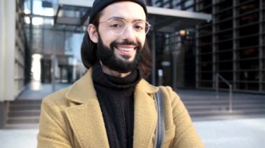 Genç adam bir ofis binasının karşısında dururken yüzünde bir gülümsemeyle kameraya bakıyor..