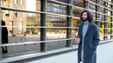 Genç hippi iş adamı gülümseyerek iş bölgesinde bir kahveyle yürüyor.