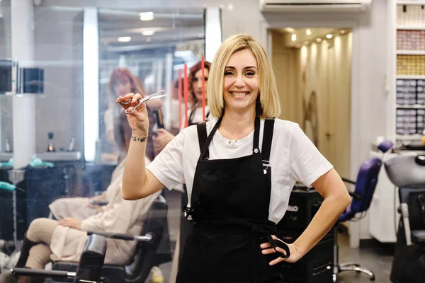 Professionell stylist tittar på kameran och ler medan du håller en sax stående i frisören. — Stockfoto