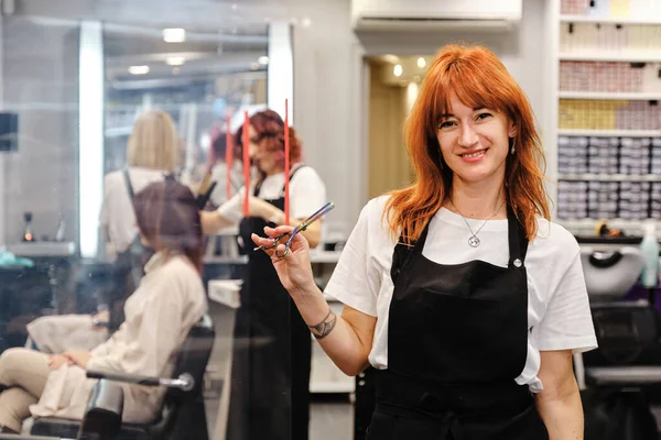 Peluquería sosteniendo una tijera en su mano mientras está de pie en el salón de belleza del cabello. —  Fotos de Stock