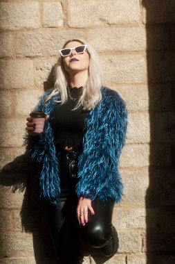 Dışarıda güneşli bir duvara yaslanırken elinde bir fincan kahve tutan rahat bir alternatif kadın..