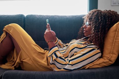 kıvırcık saçlı siyah kadın cep telefonuyla evde çalışıyor.