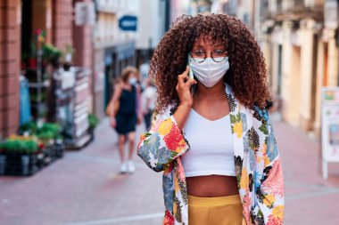 Genç kıvırcık saçlı siyah kadın sokaklarda yüz maskeli cep telefonu kullanıyor.