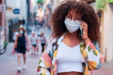 Genç kıvırcık saçlı siyah kadın sokaklarda yüz maskeli cep telefonu kullanıyor.
