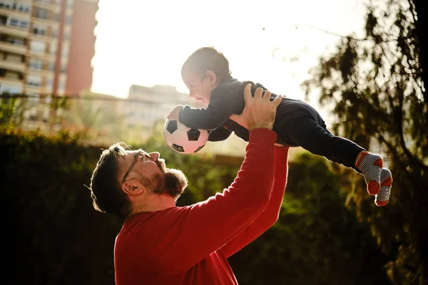 Muž se dobře baví se svým synem, zatímco ho drží ve vzduchu s fotbalovým míčem. — Stock fotografie