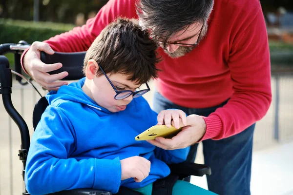 Funktionshindrade barn på rullstol och hans far använder mobiltelefon tillsammans under en promenad utomhus. — Stockfoto