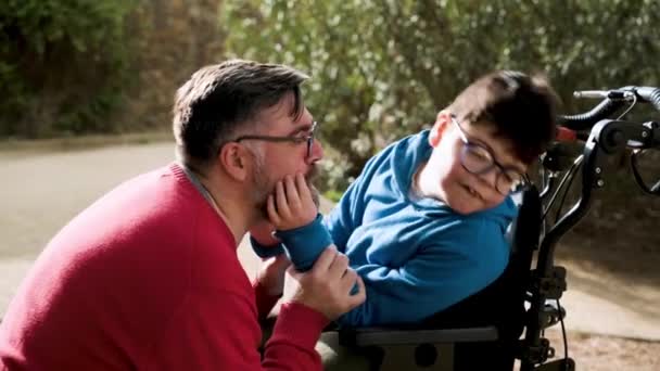 Отец развлекается со своим сыном-инвалидом в инвалидном кресле, щекоча его, пока они проводят время вместе на улице. — стоковое видео