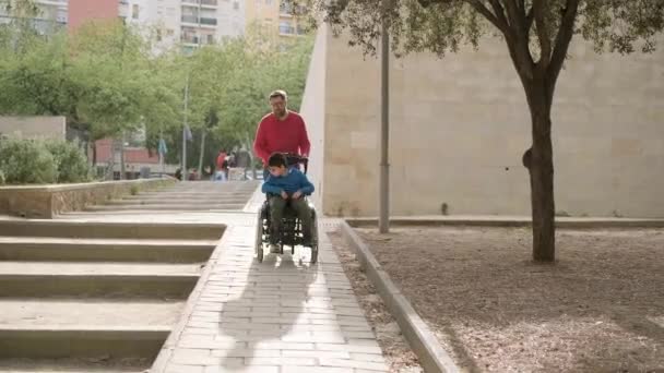 Padre spingendo la sedia a rotelle di suo figlio con tetraplegia durante una passeggiata insieme all'aperto. — Video Stock