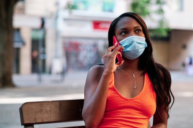 Yüz maskesi takan bir kadın telefonda konuşurken dışarıda bir bankta oturuyor..