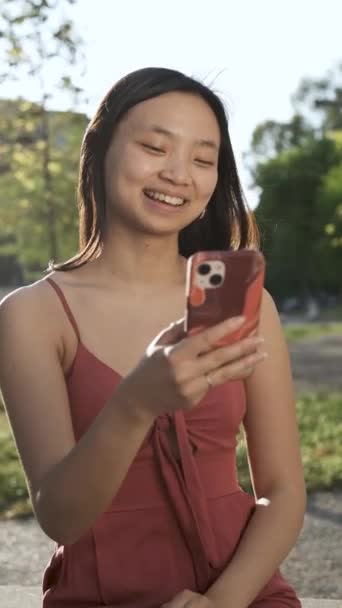 Jeune femme asiatique souriant tout en utilisant son téléphone portable à l'extérieur dans un parc. — Video