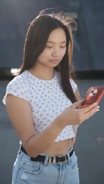 Joven mujer asiática sonriendo mientras usa su teléfono móvil al aire libre. — Vídeos de Stock